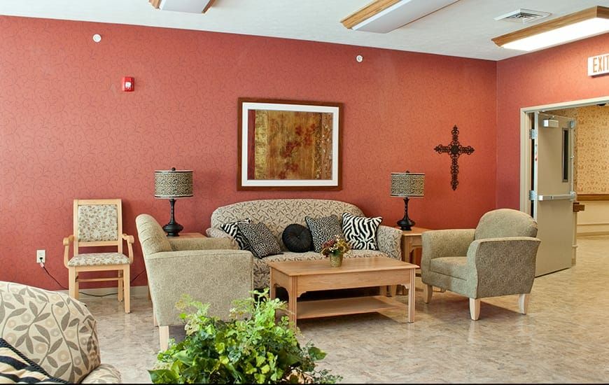 A cozy living room with a sofa and armchairs arranged around a wooden coffee table, complemented by wall art, a cross decoration, and green indoor plants.