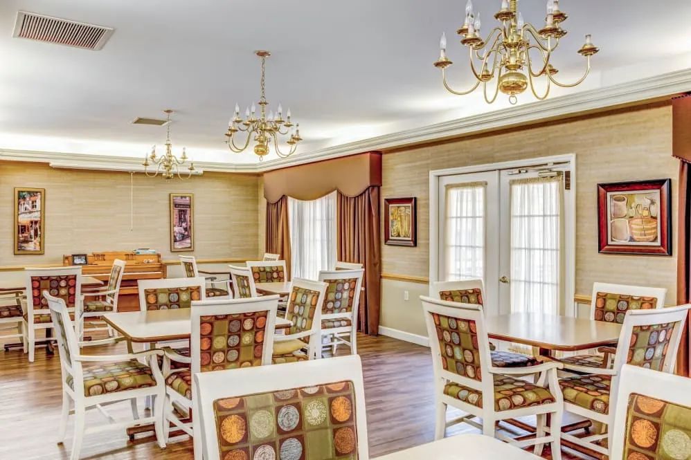 A well-lit dining room with elegant chandeliers, patterned chairs, and tables, featuring large windows with curtains and wood flooring.