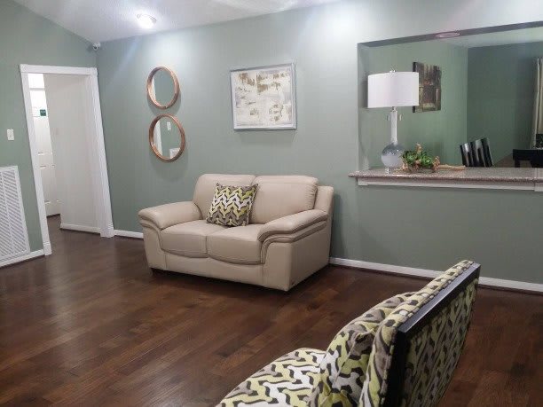 A cozy living room featuring a beige sofa and an armchair with patterned cushions, set on a wooden floor. The wall is adorned with a mirror and decorative items.