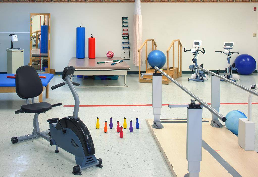 A well-equipped rehabilitation room with various exercise equipment, including an exercise bike, parallel bars, and therapy mats.