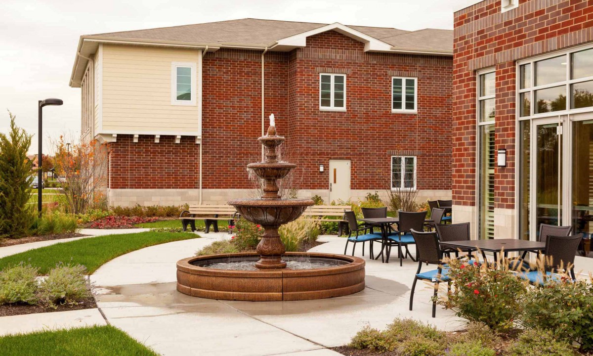 A courtyard with a central fountain surrounded by seating and a brick building in the background.