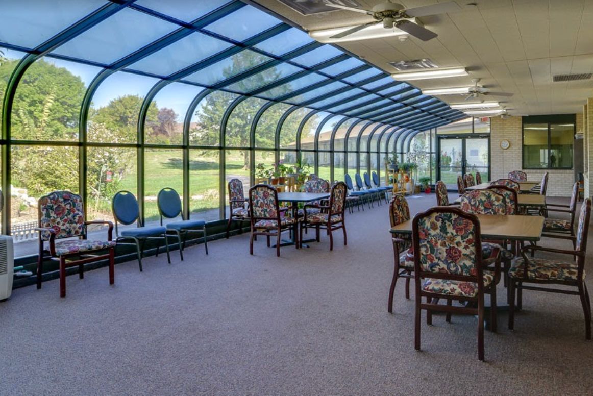 A sunlit conservatory with tables and chairs overlooking a green outdoor area.