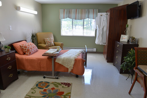 A cozy and well-lit hospital room designed for patient comfort, featuring an orange bedspread, a chair, and a window that brings in natural light.