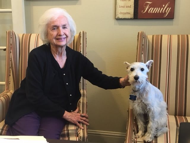 An elderly woman smiling while sitting next to a small dog indoors.