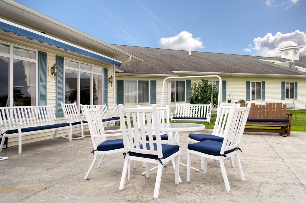 An inviting outdoor patio area featuring white chairs with blue cushions, a matching table, and a swing set against a house backdrop.