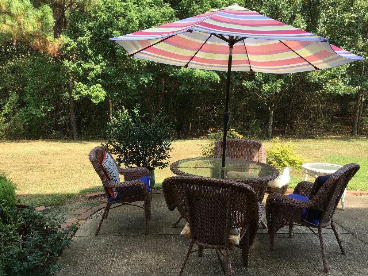 A cozy patio setup with a round table, cushioned chairs, and a colorful striped umbrella in a lush backyard.