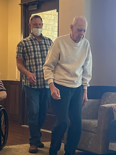 An elderly man assisted by a caregiver as they walk indoors.
