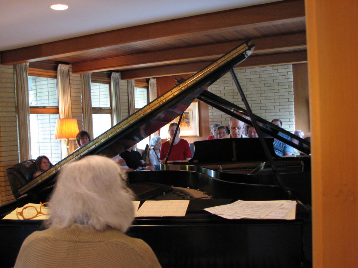 A group of people gathered around a grand piano inside a cozy room.