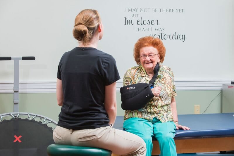 An elderly woman with an arm sling smiles while speaking with a caregiver during a physical therapy session.