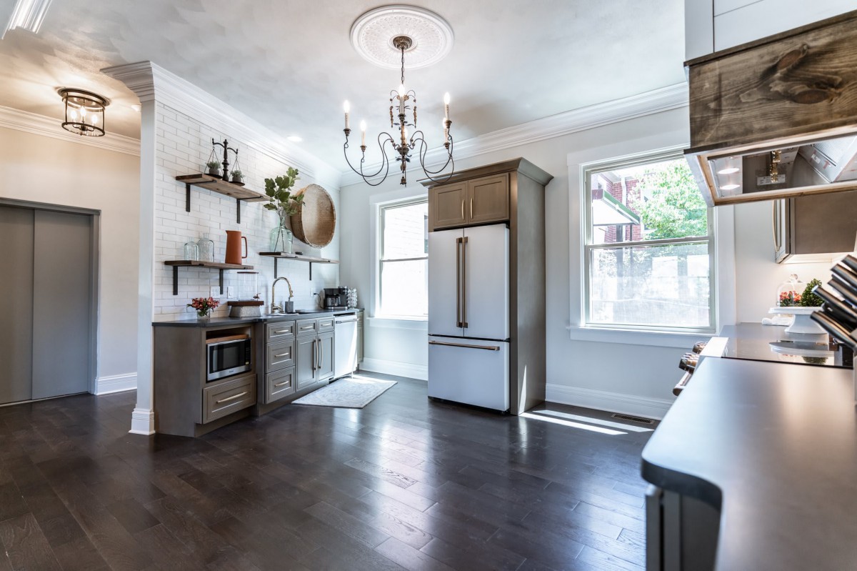 A modern kitchen with a chandelier, featuring wood flooring, open shelving, and abundant natural light.