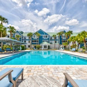 A tropical resort featuring a large swimming pool under a bright blue sky, surrounded by palm trees.