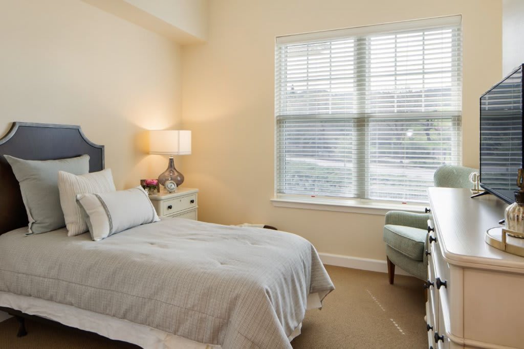 A cozy bedroom with a neatly made bed, bedside lamp, and a window letting in natural light.