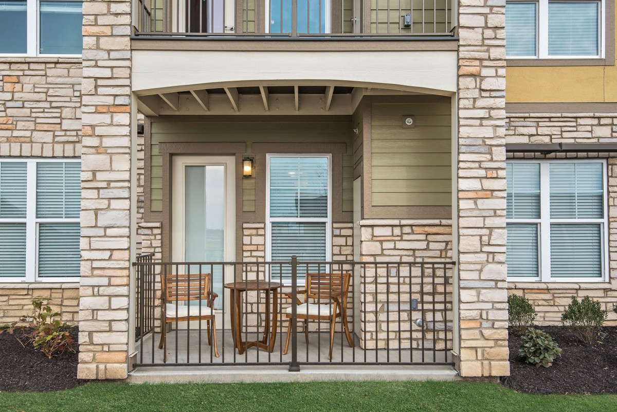 A cozy balcony with wicker chairs and a small table, enclosed by metal railings against a stone facade.