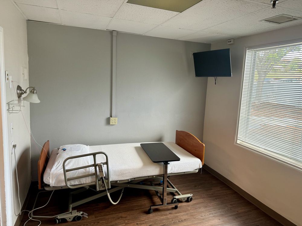 An empty hospital room with a single bed, a television on the wall, and a window providing natural light.