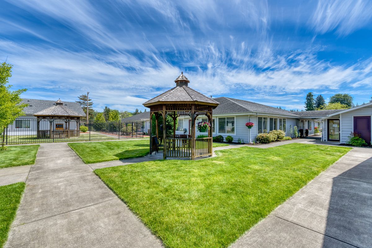 A picturesque gazebo stands in the center of a well-kept lawn, surrounded by pathways and residential buildings under a partly cloudy sky.