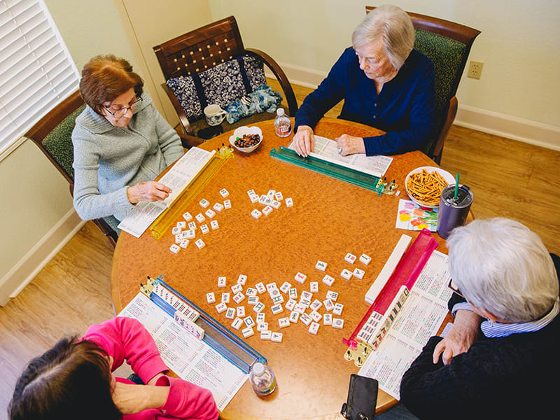 A group of seniors playing a tile-based game around a table indoors.