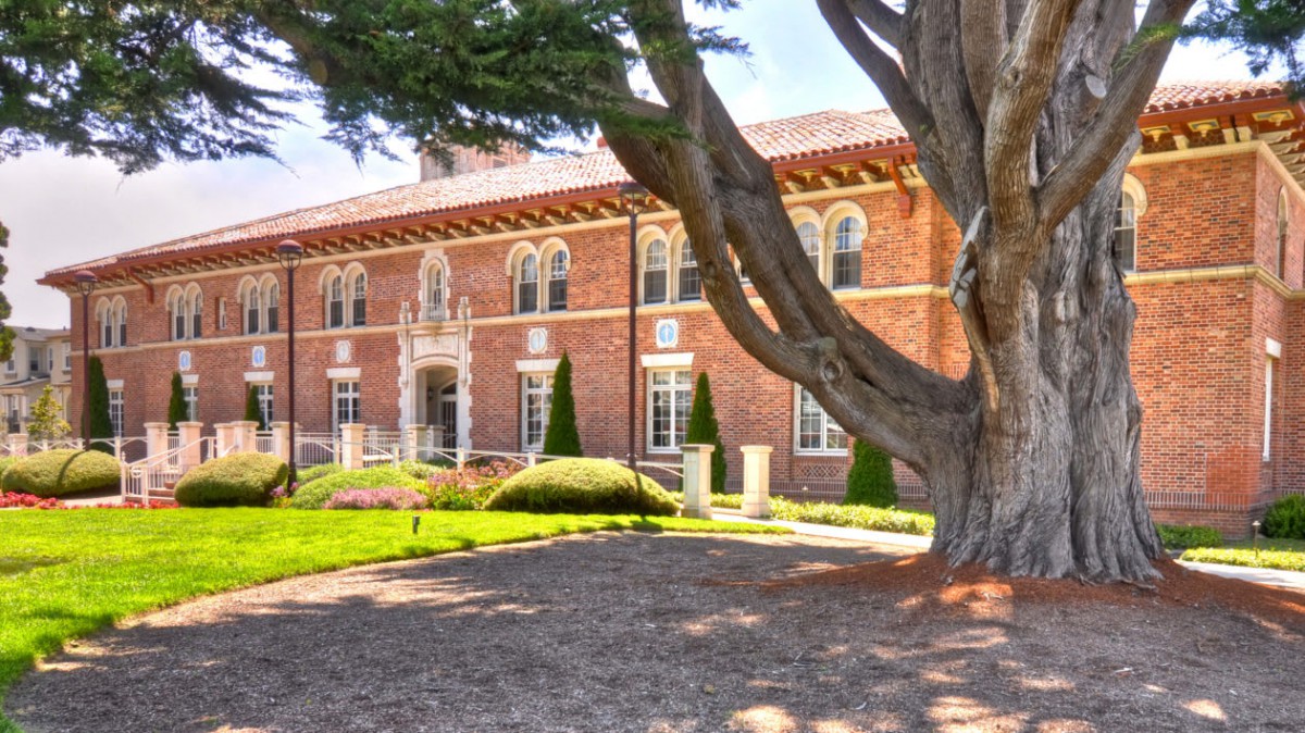 A historic brick building with lush landscaping and a large tree in the foreground.
