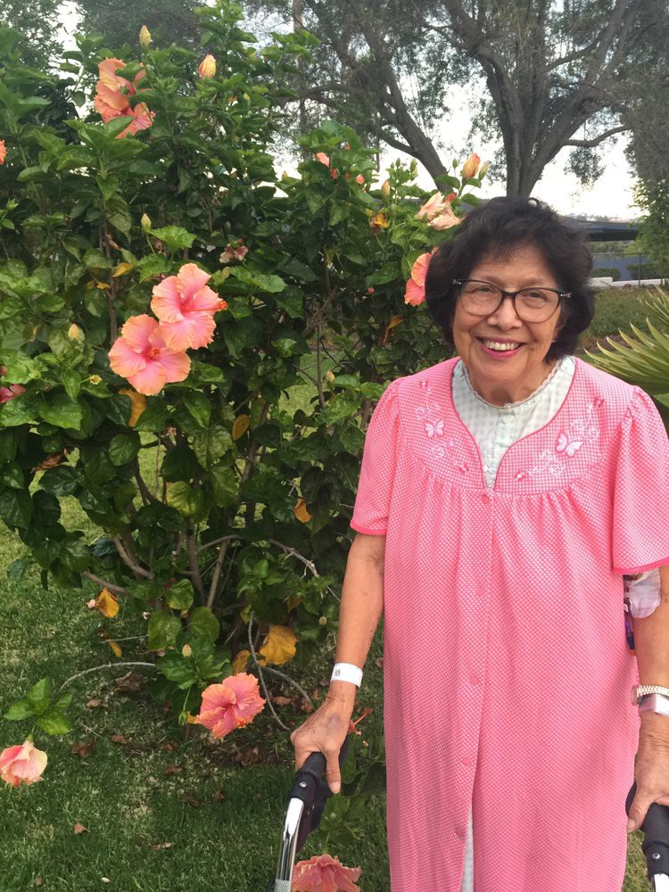 A woman in a pink dress smiles while standing next to a hibiscus bush in a garden.