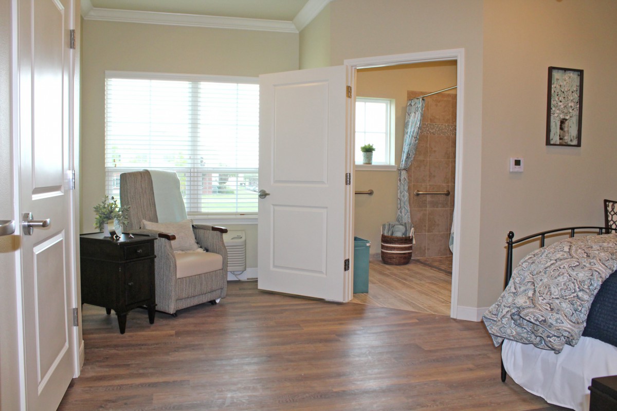 A cozy living room with a chair, open door leading to a bathroom, and natural light from a large window.