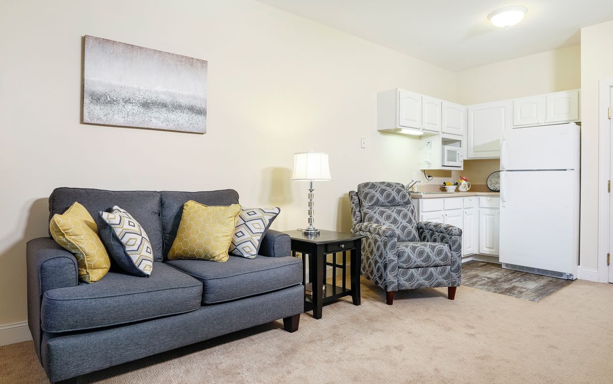 A cozy living room with a gray sofa, patterned armchair, and kitchenette in the background.