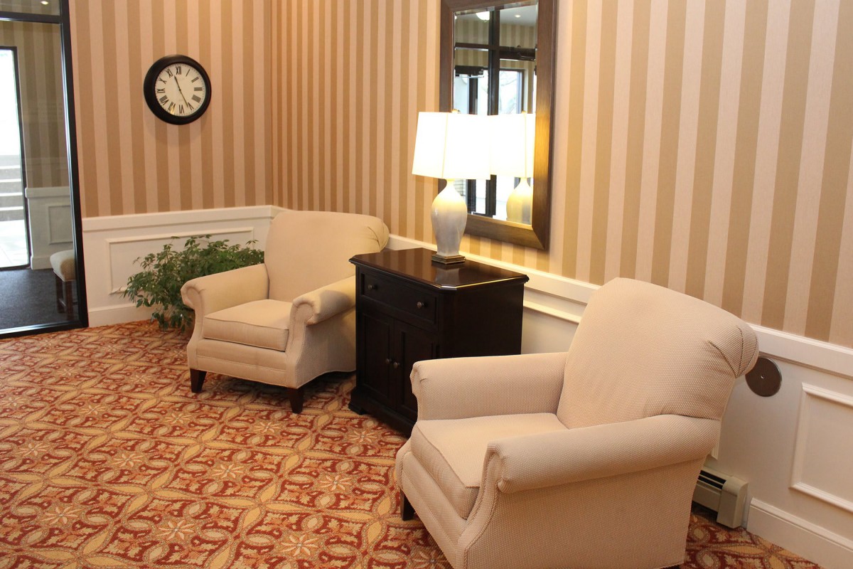 A cozy living room with two armchairs and a lamp on a side table, featuring striped wallpaper and a patterned carpet.