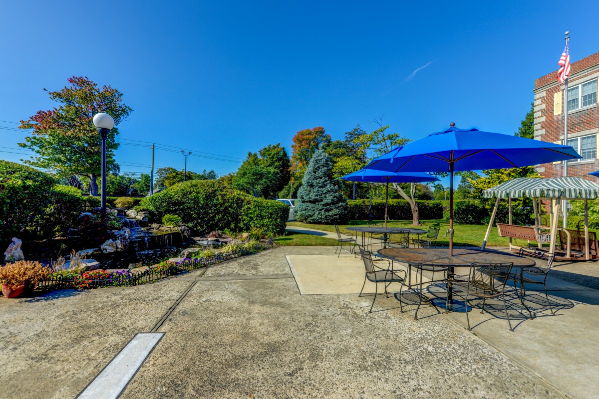 A sunny patio with outdoor furniture and a blue umbrella, surrounded by a landscaped garden.