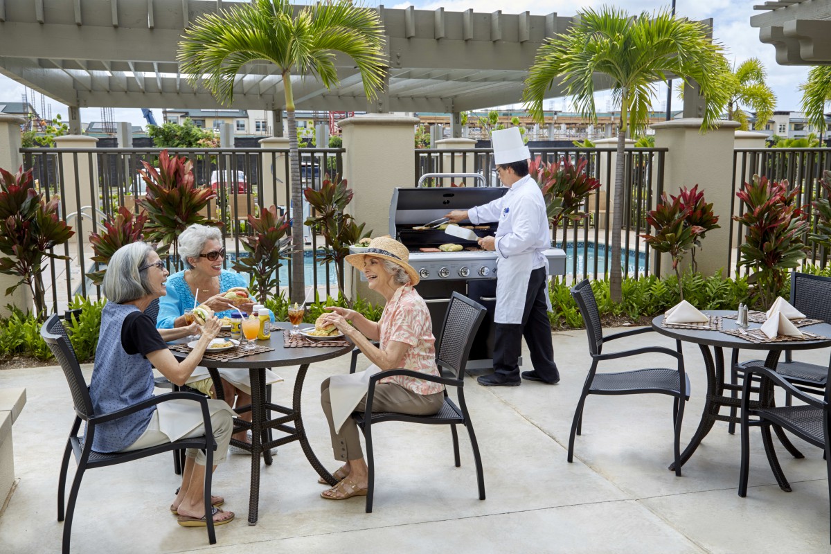 A group of people enjoying a meal outdoors while a chef cooks on a barbecue grill by the poolside.
