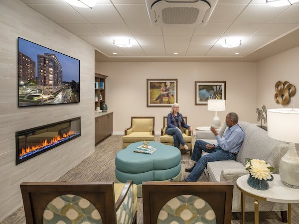A cozy lounge area with two people having a conversation, featuring comfortable seating, a fireplace, and a wall-mounted television.