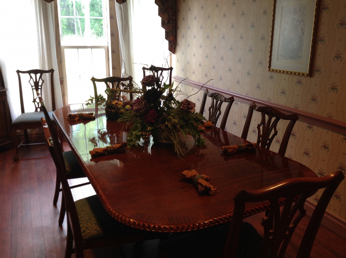 A dining room with a large wooden table and chairs, decorated with a floral centerpiece near a window.