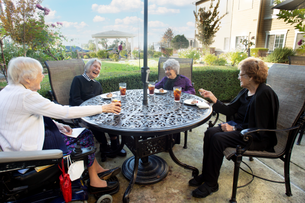 A group of elderly women enjoying drinks and conversation at an outdoor table in a garden setting.