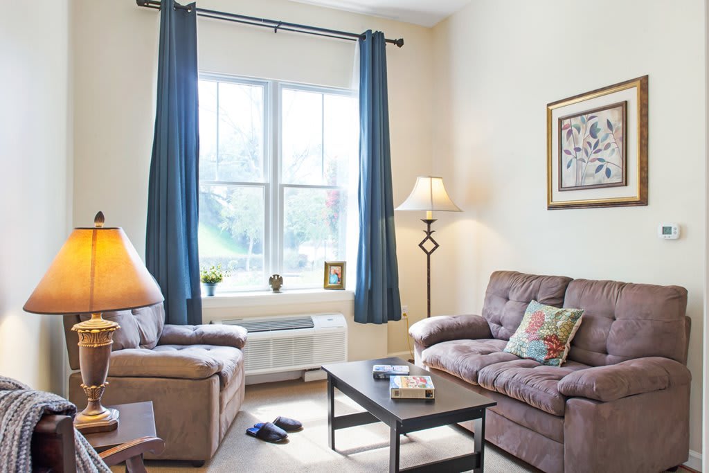 A cozy living room with a sofa, lamp, coffee table, and large window framed by blue curtains.