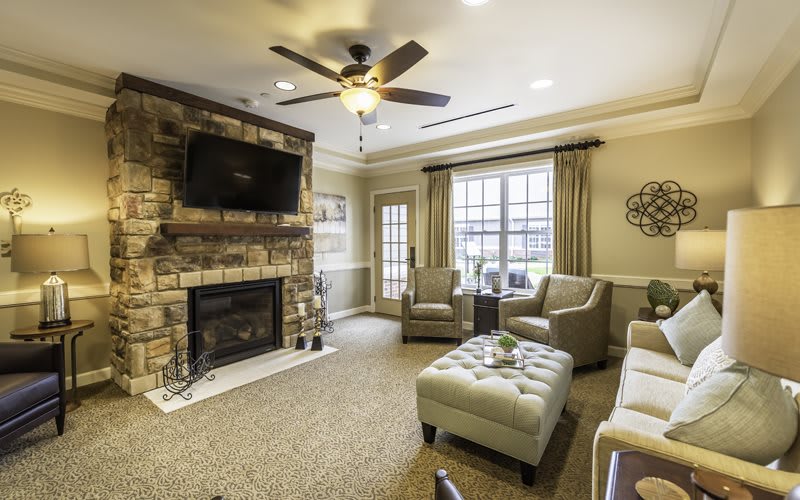A cozy living room with comfortable seating and a stone fireplace, featuring a ceiling fan and large windows.