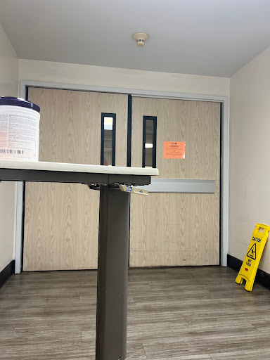 A waiting area featuring double doors, a caution sign on the floor, and a container of cleaning supplies on a table.