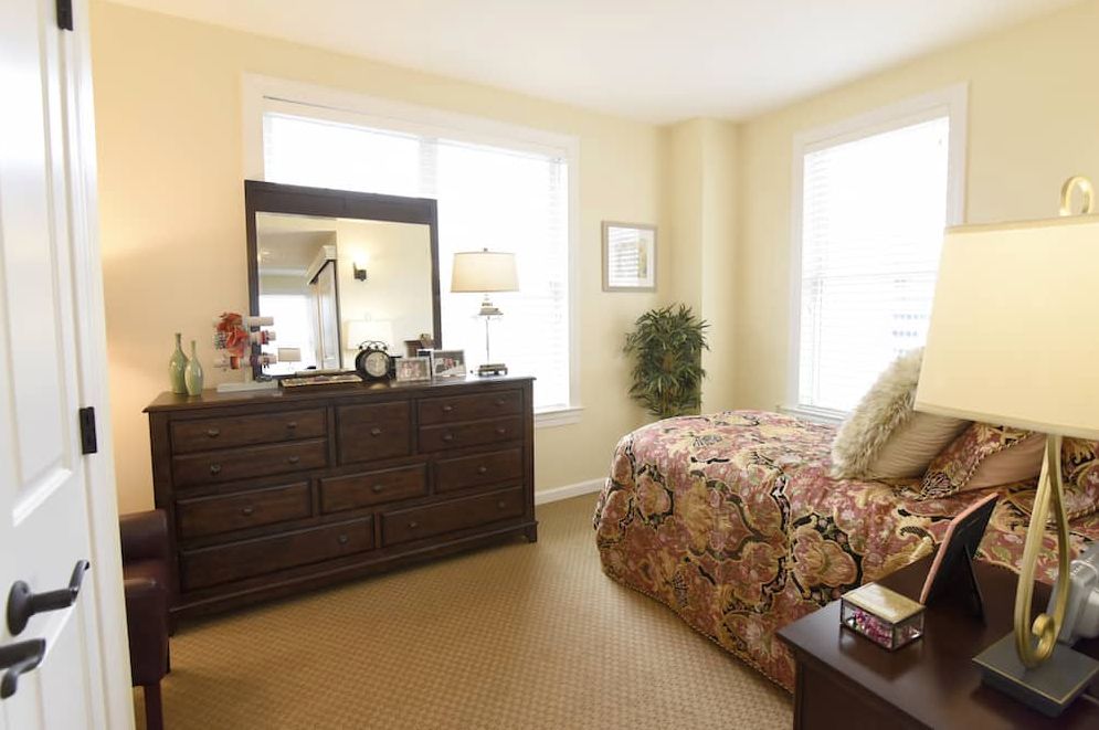 A cozy bedroom featuring a bed with a floral comforter, a wooden dresser with a mirror, and natural light streaming through the windows.