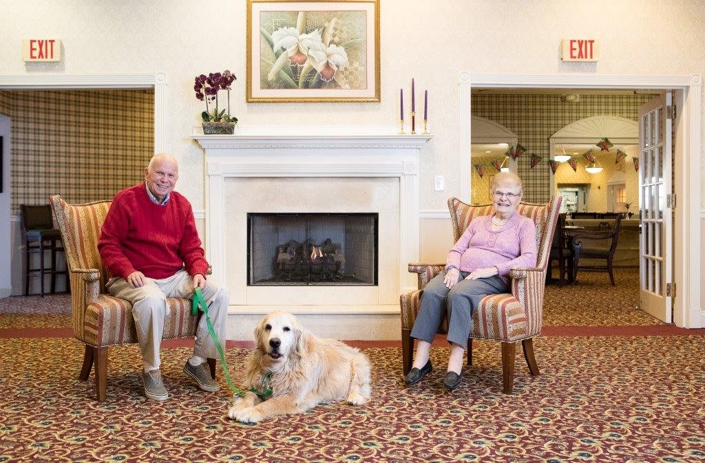 An elderly couple sitting by a fireplace with their dog in a comfortable living room.