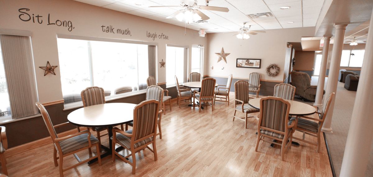 A cozy dining room with wooden floors, tables, and chairs, featuring ceiling fans and large windows for natural light.