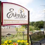 A sign for Eventide Senior Living Community with flowers and a bench nearby.