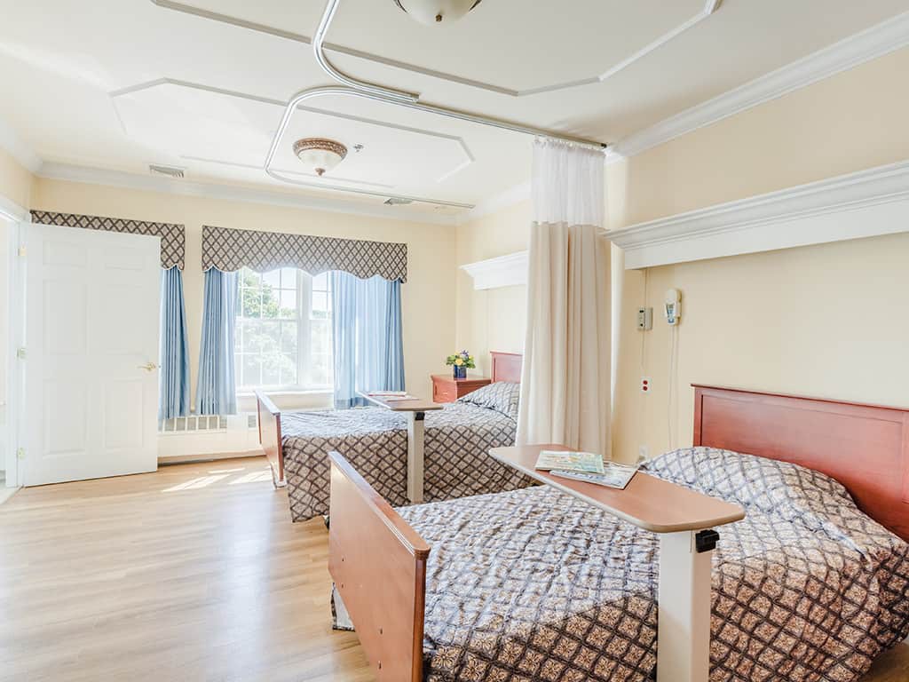 An empty hospital room with two beds and large windows letting in natural light.