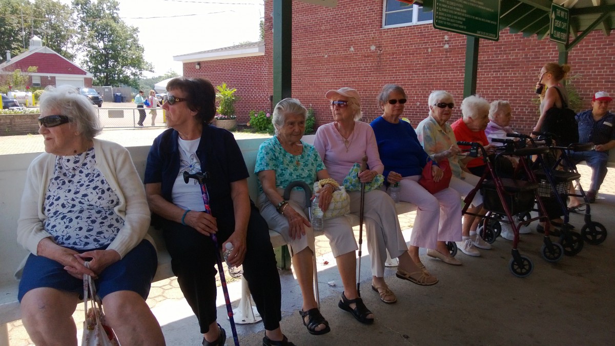 A group of elderly women sitting on a bench outdoors on a sunny day.