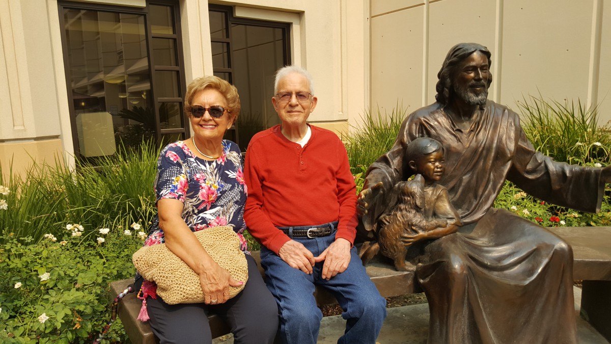 An elderly couple sitting on a bench next to a sculpture, enjoying a sunny day.