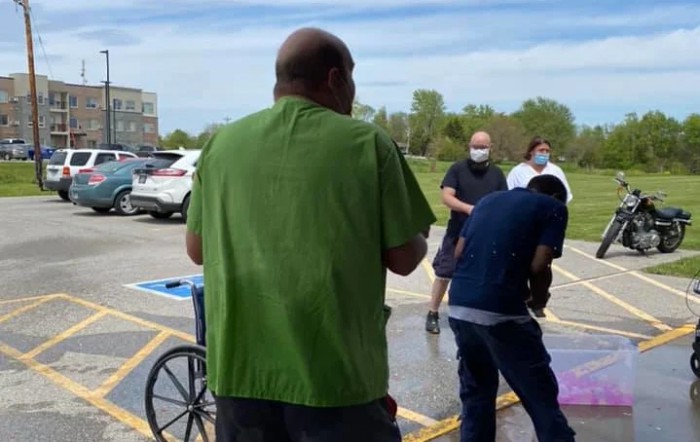 People gathered in a parking lot, some wearing masks, with a wheelchair visible.