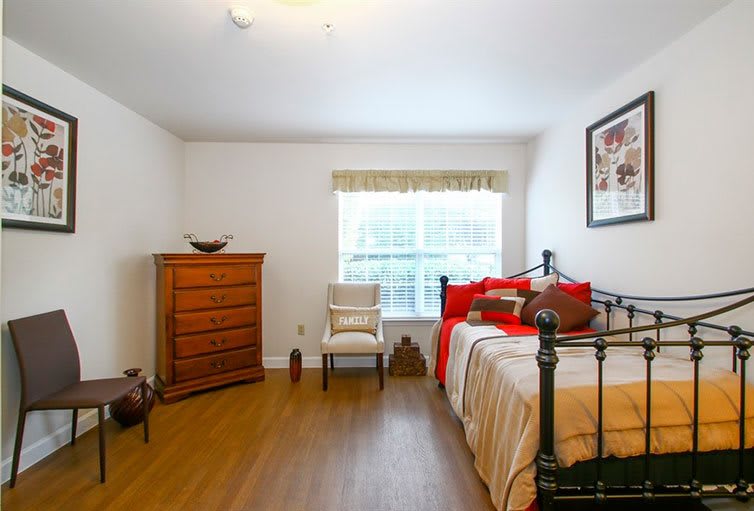 A cozy bedroom with wooden flooring, featuring a single bed with red and beige bedding, a wooden dresser, and a chair by the window that allows natural light to fill the room.