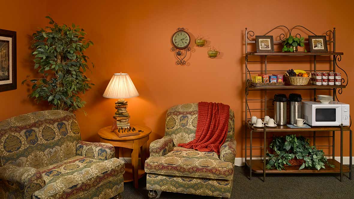 A cozy living room with an orange wall, featuring patterned armchairs, a lamp, and a decorative bookshelf.