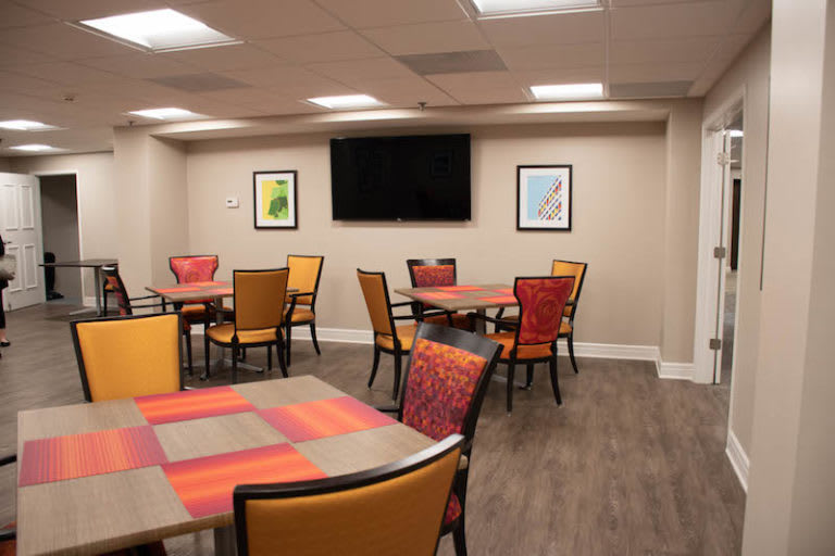 A modern dining area featuring colorful tables and chairs, with framed artwork on the walls and a television mounted above.