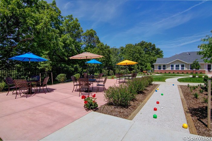 A serene outdoor patio with tables, umbrellas, and a bocce court surrounded by lush greenery.
