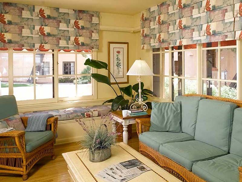 A cozy living room featuring a wicker sofa set with light green cushions, a wooden coffee table, large windows with patterned curtains, and decorative plants.