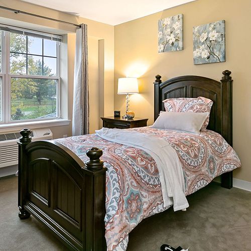 A cozy bedroom with a single bed, adorned with floral bedding and a soft throw, beside a window and a nightstand with a lamp.