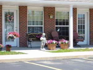 A cozy patio area in front of a brick building with benches, chairs, flower pots, and a small statue.