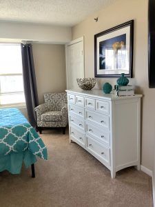 A cozy bedroom featuring a white dresser, an upholstered armchair, and a bed with teal bedding.