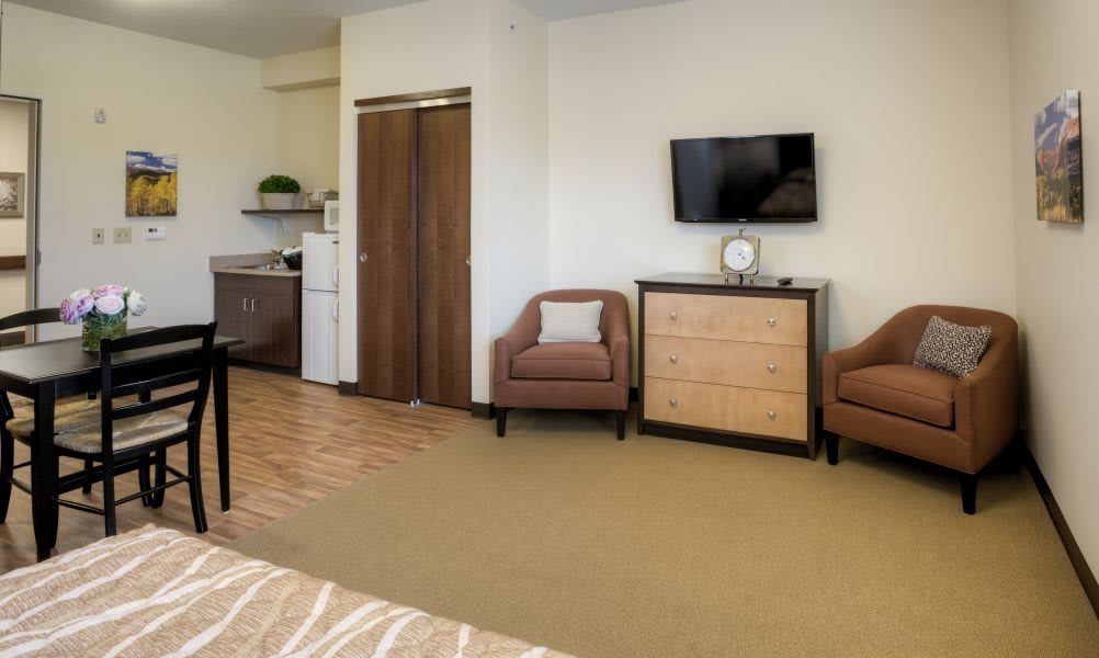 A spacious hotel room featuring a kitchenette, two armchairs, a dresser, and a wall-mounted TV.